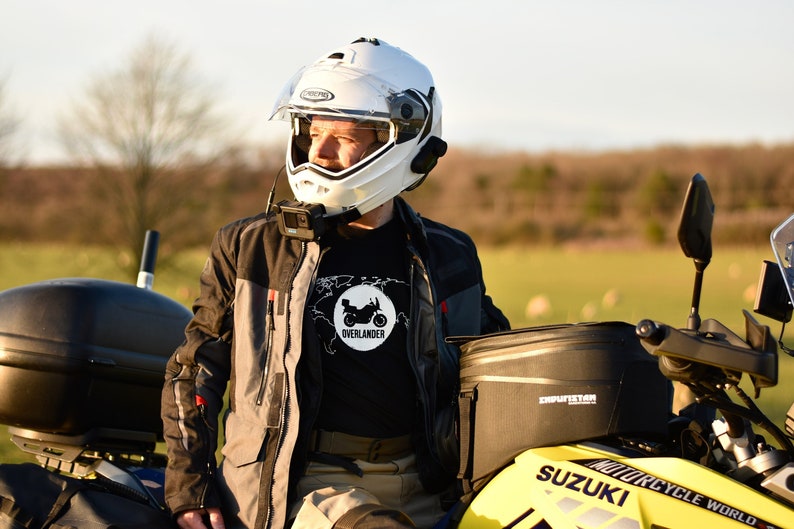 Op de afbeelding: Een persoon met een witte helm en een zwart T-shirt met een motorfietsafbeelding en de tekst "Overlander" zit op een gele Suzuki motorfiets. De motorfiets heeft zwarte zadeltassen en een zwarte topkoffer.