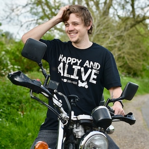 May include: A man wearing a black t-shirt with white text that reads "Happy and Alive" is sitting on a silver motorcycle. He is smiling and has his hand on his head.