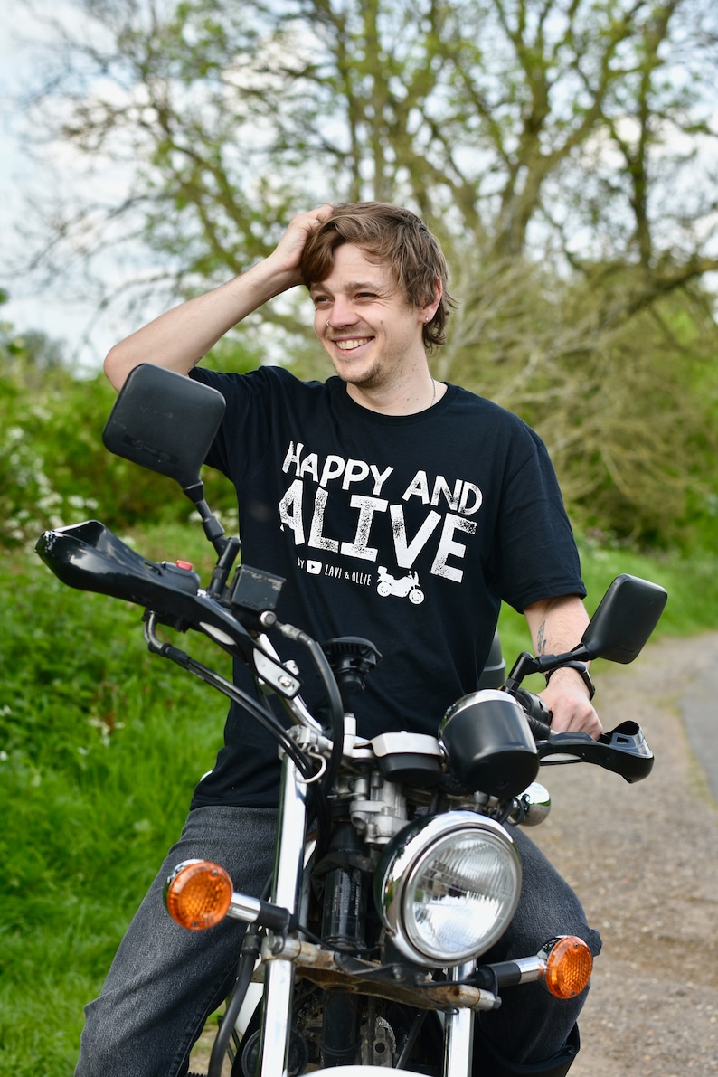 May include: A man wearing a black t-shirt with white text that reads "Happy and Alive" is sitting on a silver motorcycle. He is smiling and has his hand on his head.