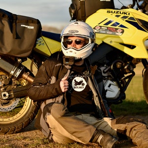 Op de afbeelding: Een persoon met een witte helm en een zwart motorjack zit naast een gele Suzuki motorfiets. De motorfiets heeft een zwart-witte sticker met de tekst "Overlander" op het jack. De motorfiets heeft een zwarte uitlaatpijp en een zwart-gouden wiel.
