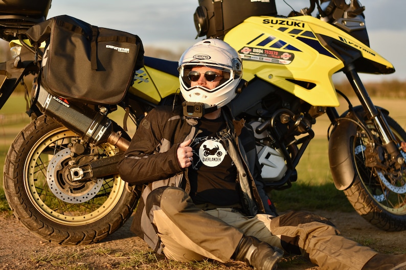 Op de afbeelding: Een persoon met een witte helm en een zwart motorjack zit naast een gele Suzuki motorfiets. De motorfiets heeft een zwart-witte sticker met de tekst "Overlander" op het jack. De motorfiets heeft een zwarte uitlaatpijp en een zwart-gouden wiel.