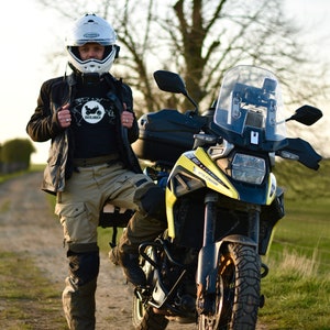Op de afbeelding: Een persoon in een zwart jack, helm en kaki broek staat naast een gele en zwarte motorfiets. De motorfiets heeft een groot windscherm en een zwarte topkoffer.