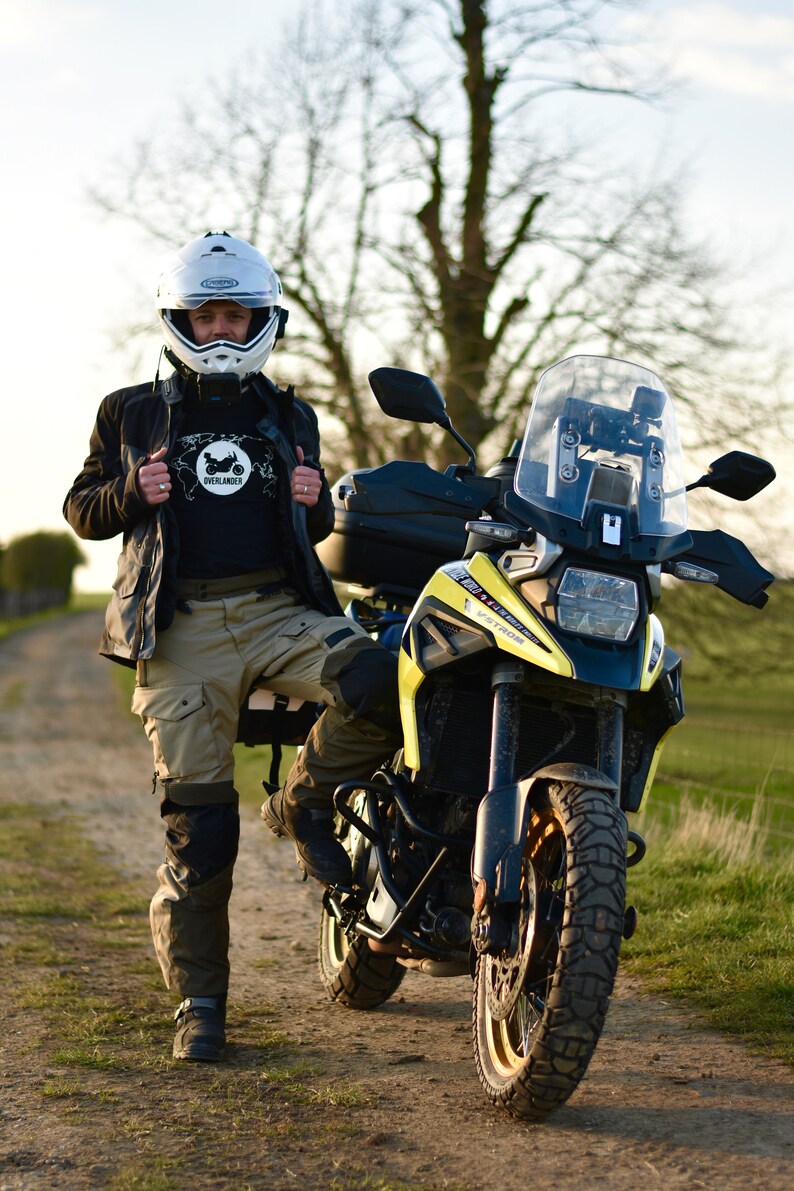 Op de afbeelding: Een persoon in een zwart jack, helm en kaki broek staat naast een gele en zwarte motorfiets. De motorfiets heeft een groot windscherm en een zwarte topkoffer.