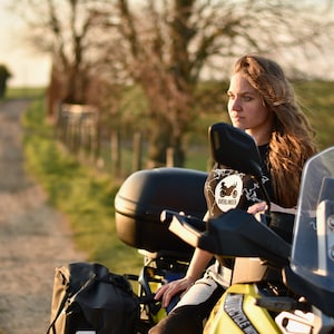 Op de afbeelding: Een vrouw zit op een gele motorfiets met zwarte bagage en een windscherm. Ze draagt een zwart T-shirt met een wit logo met de tekst "Overlander" en een motorfietsafbeelding. Ze kijkt naar de zijkant.
