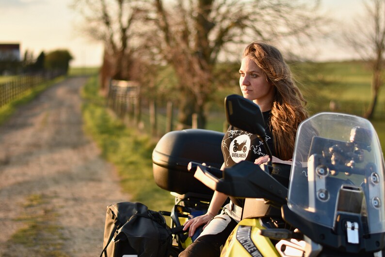 Op de afbeelding: Een vrouw zit op een gele motorfiets met zwarte bagage en een windscherm. Ze draagt een zwart T-shirt met een wit logo met de tekst "Overlander" en een motorfietsafbeelding. Ze kijkt naar de zijkant.