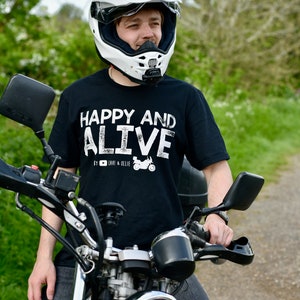 May include: A person wearing a black t-shirt with the text "HAPPY AND ALIVE" and a white motorcycle helmet, sitting on a black motorcycle. The motorcycle has a chrome headlight and handlebars.