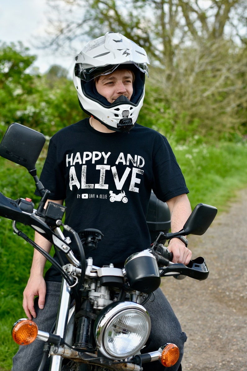 May include: A person wearing a black t-shirt with the text "HAPPY AND ALIVE" and a white motorcycle helmet, sitting on a black motorcycle. The motorcycle has a chrome headlight and handlebars.