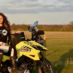 Op de afbeelding: Een vrouw in een zwart T-shirt met een motorfietsafbeelding staat naast een gele Suzuki-motorfiets. De motorfiets heeft zwarte bagagezakken en een zwart windscherm. De vrouw draagt zwarte motorlaarzen en een wit-zwarte motorjas.