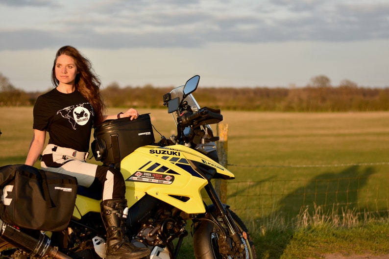 Op de afbeelding: Een vrouw in een zwart T-shirt met een motorfietsafbeelding staat naast een gele Suzuki-motorfiets. De motorfiets heeft zwarte bagagezakken en een zwart windscherm. De vrouw draagt zwarte motorlaarzen en een wit-zwarte motorjas.