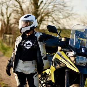 Op de afbeelding: Een persoon met een witte helm en een zwart T-shirt met een witte afbeelding van een motorfiets en het woord "Overlander" staat naast een gele en zwarte motorfiets. De motorfiets heeft een helder windscherm en een zwarte stoel.