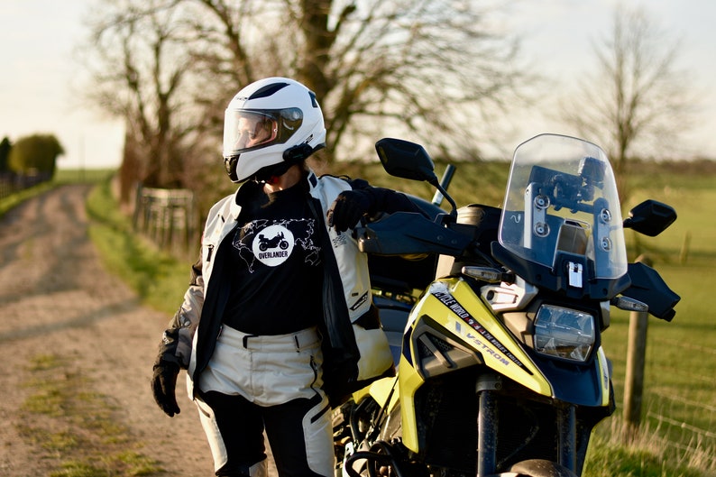 Op de afbeelding: Een persoon met een witte helm en een zwart T-shirt met een witte afbeelding van een motorfiets en het woord "Overlander" staat naast een gele en zwarte motorfiets. De motorfiets heeft een helder windscherm en een zwarte stoel.