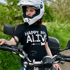 May include: A person wearing a white motorcycle helmet and a black t-shirt with the text "Happy and Alive" in white. The person is sitting on a motorcycle with a black seat and a chrome frame.