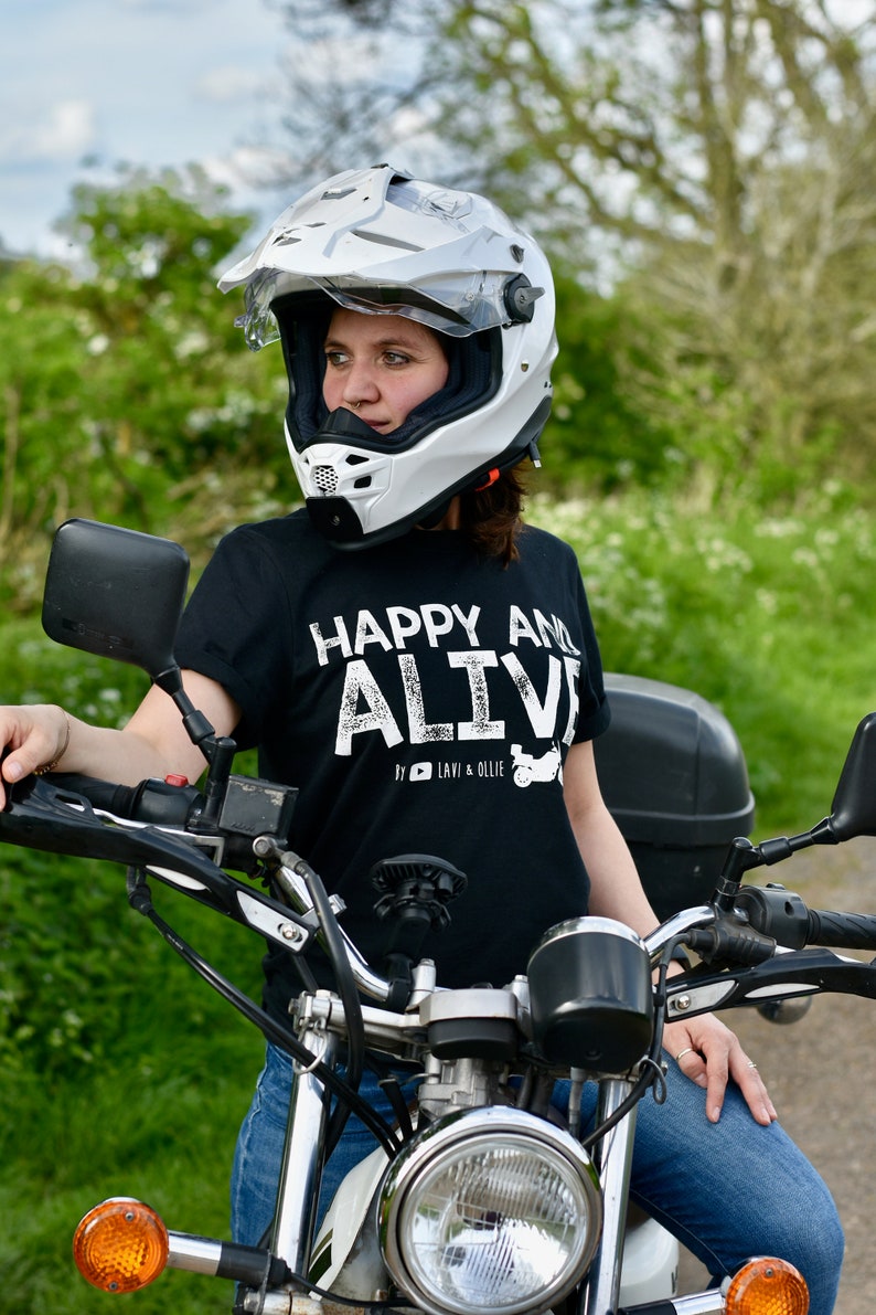 May include: A person wearing a white motorcycle helmet and a black t-shirt with the text "Happy and Alive" in white. The person is sitting on a motorcycle with a black seat and a chrome frame.