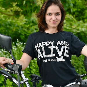 May include: A woman wearing a black t-shirt with white text that reads "Happy and Alive" and a motorcycle graphic. She is sitting on a motorcycle with a green background.