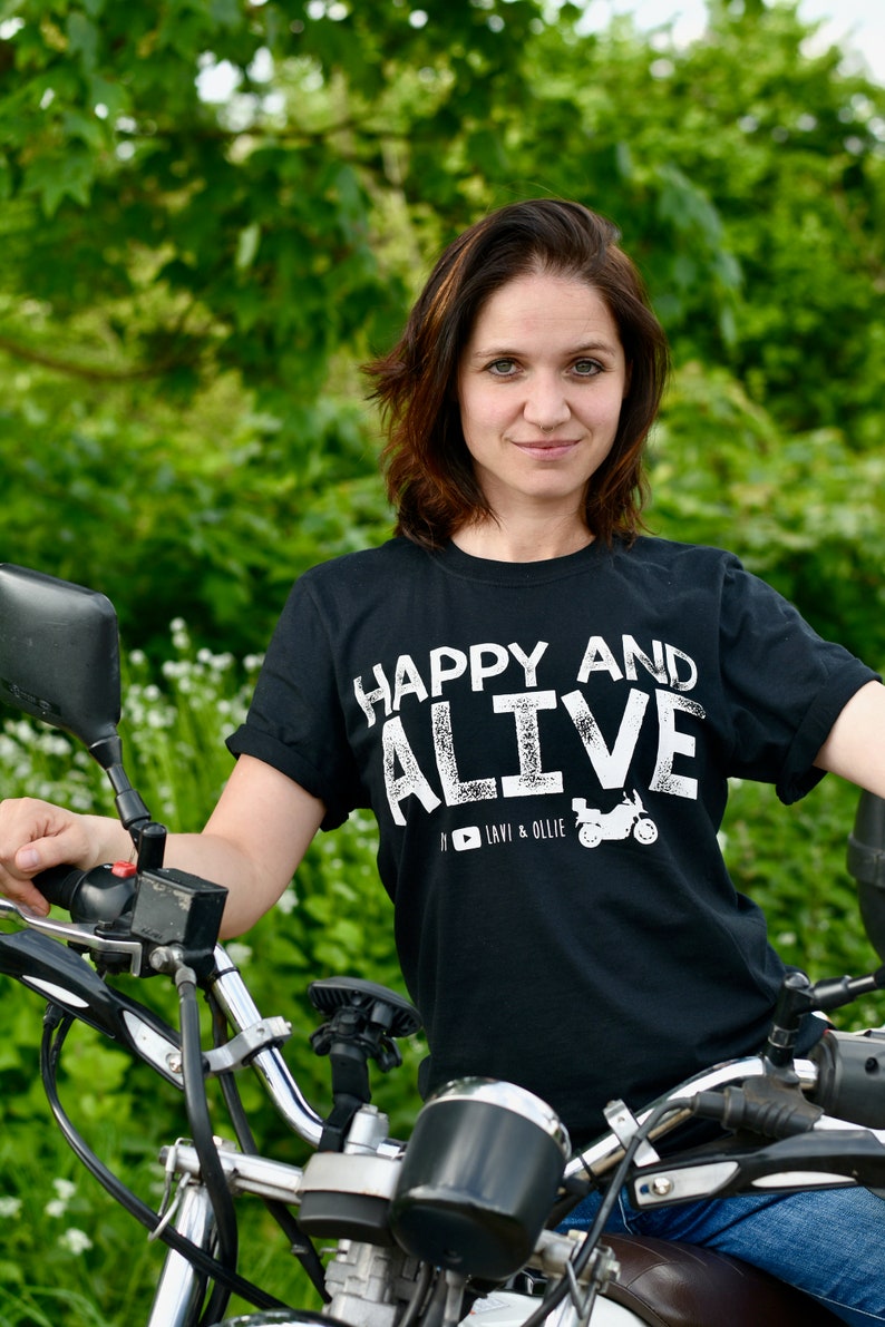 May include: A woman wearing a black t-shirt with white text that reads "Happy and Alive" and a motorcycle graphic. She is sitting on a motorcycle with a green background.