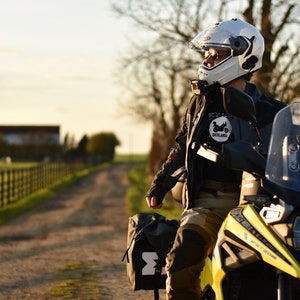 Op de afbeelding: Een persoon met een witte helm en een zwart motorjack zit op een gele en zwarte motor. De motor heeft een windscherm en zadeltassen. De persoon kijkt naar rechts, en de achtergrond is een landelijke omgeving met een hek en een onverharde weg.