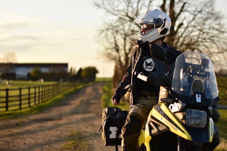 Op de afbeelding: Een persoon met een witte helm en een zwart motorjack zit op een gele en zwarte motor. De motor heeft een windscherm en zadeltassen. De persoon kijkt naar rechts, en de achtergrond is een landelijke omgeving met een hek en een onverharde weg.