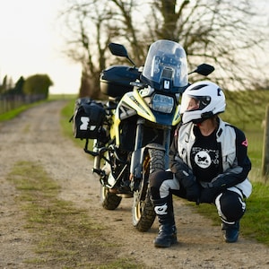 Op de afbeelding: Een persoon met een witte helm en motorfietskleding zit naast een gele en zwarte motorfiets op een onverharde weg. De motorfiets heeft zadeltassen en een windscherm.