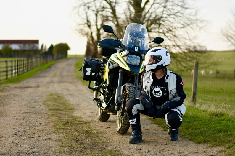 Op de afbeelding: Een persoon met een witte helm en motorfietskleding zit naast een gele en zwarte motorfiets op een onverharde weg. De motorfiets heeft zadeltassen en een windscherm.