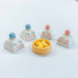 May include: Four white ceramic cat-shaped keycaps with blue, pink, and white decorative accents. A small yellow bowl with four yellow dumplings sits in the center.