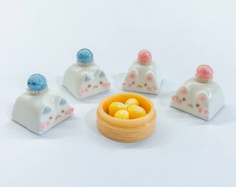 May include: Four white ceramic cat-shaped keycaps with blue, pink, and white decorative accents. A small yellow bowl with four yellow dumplings sits in the center.