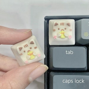 May include: Two white keycaps featuring a cute cartoon cat design holding a yellow object. The keycaps are on a black keyboard with the keys "tab" and "caps lock" visible.