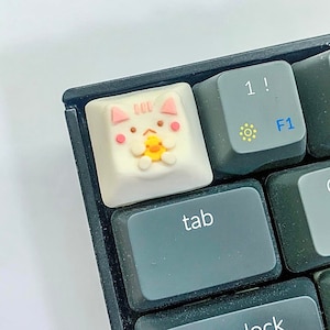 May include: A white keyboard keycap with a pink and white cat design holding a yellow chick. The keycap is labeled "tab" and is on a black keyboard.