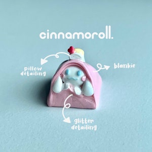 May include: A pink and white Cinnamoroll character figurine, with a pink blanket and a white pillow. The figurine has blue eyes and a pink nose. The figurine is covered in glitter.