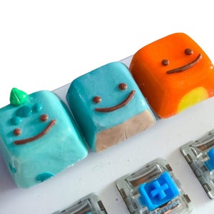 May include: Three custom keycaps with smiling faces. The keycaps are blue, orange, and green. The blue keycap has a small green leaf on top. The orange keycap has a yellow accent. The keycaps are on a white keyboard.