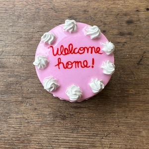 May include: A pink, round cake with white frosting swirls and the words "welcome home!" in red script. The cake is a decorative item, possibly a figurine or a small serving piece.