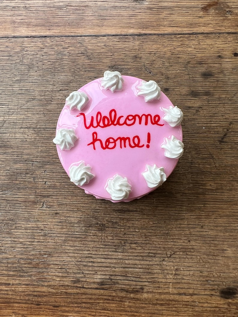 May include: A pink, round cake with white frosting swirls and the words "welcome home!" in red script. The cake is a decorative item, possibly a figurine or a small serving piece.