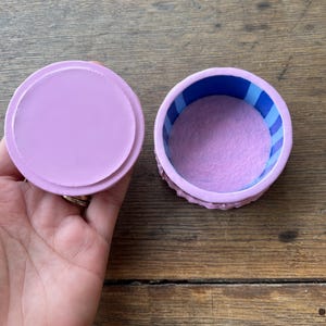 May include: A small, round, open jewelry box with a lid. The box is lavender with a blue and white striped interior. The inside of the box is lined with a soft, lavender material. The lid is a solid lavender color.