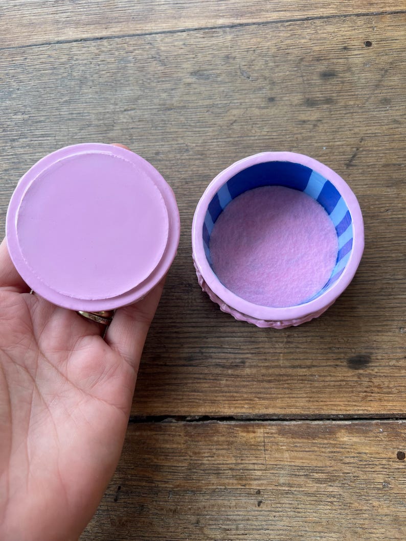 May include: A small, round, open jewelry box with a lid. The box is lavender with a blue and white striped interior. The inside of the box is lined with a soft, lavender material. The lid is a solid lavender color.