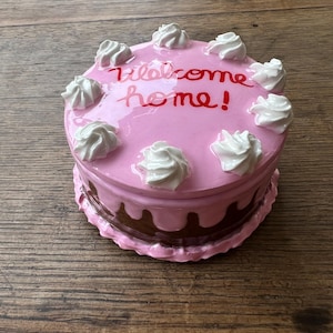 May include: A pink ceramic cake-shaped box with white frosting swirls and the words "Welcome home!" in red script. The bottom layer is brown with a dripping effect, and the box sits on a wooden surface.