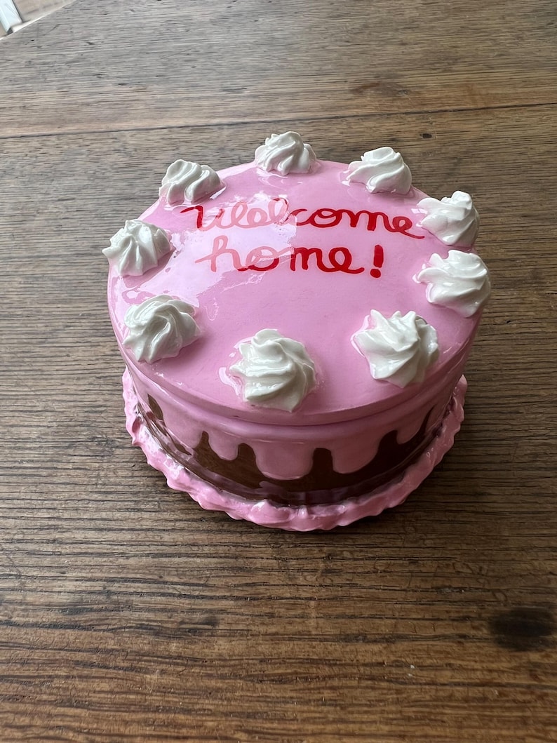 May include: A pink ceramic cake-shaped box with white frosting swirls and the words "Welcome home!" in red script. The bottom layer is brown with a dripping effect, and the box sits on a wooden surface.