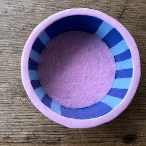 May include: A small, round, ceramic bowl with a lavender exterior and a blue and light blue striped interior. The bowl's interior features a soft, light purple felt lining. The bowl is sitting on a wooden surface.