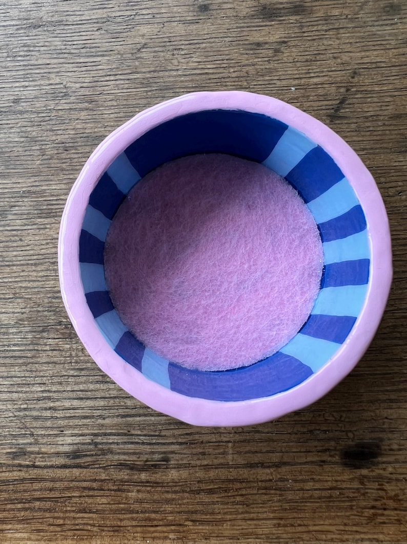 May include: A small, round, ceramic bowl with a lavender exterior and a blue and light blue striped interior. The bowl's interior features a soft, light purple felt lining. The bowl is sitting on a wooden surface.