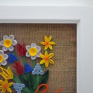 May include: A framed paper flower bouquet in a white frame. The flowers are crafted from paper in white, yellow, red, blue, and green, set against a burlap backdrop. A decorative piece.