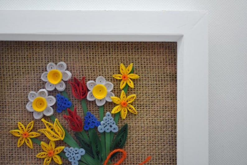 May include: A framed paper flower bouquet in a white frame. The flowers are crafted from paper in white, yellow, red, blue, and green, set against a burlap backdrop. A decorative piece.