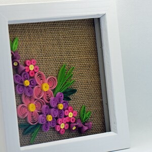Pu&ograve; includere: Un'opera d'arte di fiori di carta incorniciata con fiori rosa, viola e gialli su uno sfondo di juta.