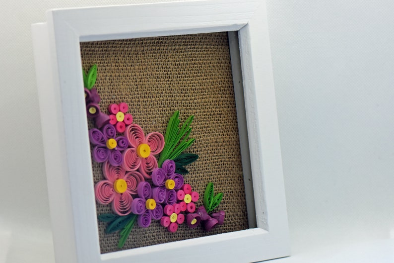 Pu&ograve; includere: Un'opera d'arte di fiori di carta incorniciata con fiori rosa, viola e gialli su uno sfondo di juta.