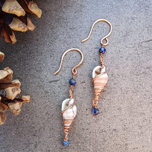 Op de afbeelding: Een paar hangende oorbellen met kleine, lichtgekleurde spiraalvormige schelpen omwikkeld met koperdraad. Elke oorbel bevat twee kleine, gefacetteerde donkerblauwe lapis lazuli kralen en een gebogen koperen haakje voor aan de oorlel.