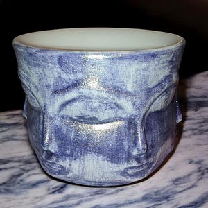 May include: A handcrafted ceramic cup with a unique design. The cup is painted in shades of blue and features the sculpted faces of three figures. The interior is white, and the cup sits on a marble surface. The cup is a decorative item.