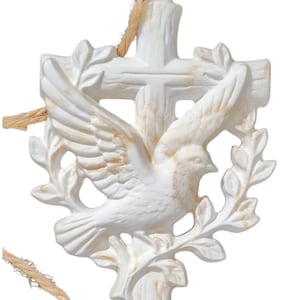 May include: A white cross-shaped ornament with a dove and olive branch design. The dove has outstretched wings and is surrounded by a wreath. The ornament is made of a light-colored material and has a natural-colored rope for hanging.