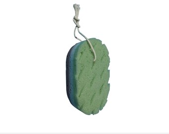May include: An oval-shaped bath sponge featuring three distinct layers of foam in light green, blue, and dark purple. The surface has a textured, diamond-patterned design and includes a white cord loop attached at the top for hanging.