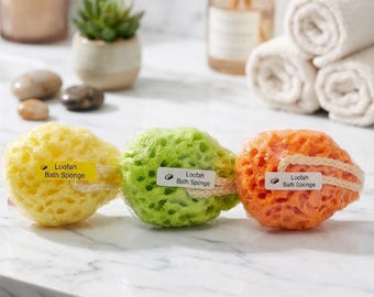 May include: Three round bath sponges in yellow, green, and orange, each featuring a textured, porous surface and a beige hanging rope. Each sponge has a small rectangular label attached that reads "Loofah Bath Sponge" with a small icon.