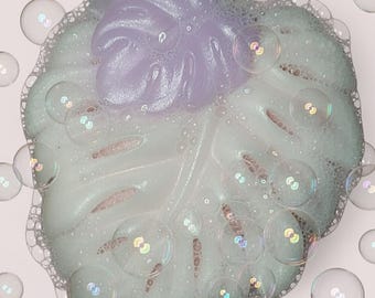May include: A leaf-shaped bar of soap with a light green base and a lavender-colored top section. The surface of the soap is covered in a layer of white lather and small bubbles, with several larger, translucent soap bubbles floating around it.