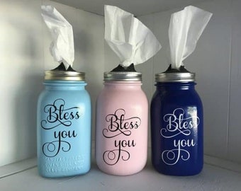 May include: Three mason jars painted in blue, pink, and dark blue with the words "Bless You" in black lettering. Each jar has a white tissue dispenser on top.