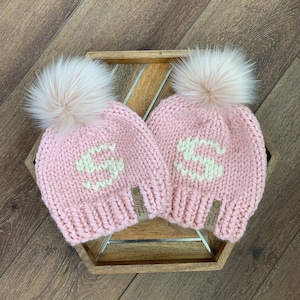 May include: Two pink knit hats with white pom poms and white knit letters. The hats have brown leather tags that say "Handknit & Made".
