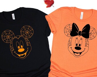 May include: Two t-shirts with a Halloween theme. One black t-shirt features Mickey Mouse with a spiderweb design. The other orange t-shirt features Minnie Mouse with a spiderweb design and a black bow.